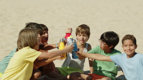 Excited-boys-sitting-on-beach-and-clinking-with-water-bottles